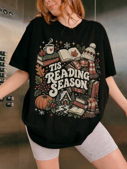 Black Comfort Colors shirt modeled with a Tis Reading Season graphic featuring books, coffee, autumn leaves, pumpkin, scarf, winter hat and snowy cottage, cozy book lover t shirt for readers