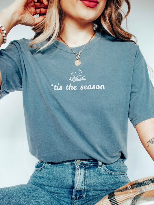 A woman wearing a blue jean crewneck t-shirt with a white graphic that reads "'tis the season" under an open book illustration, paired with blue jeans.