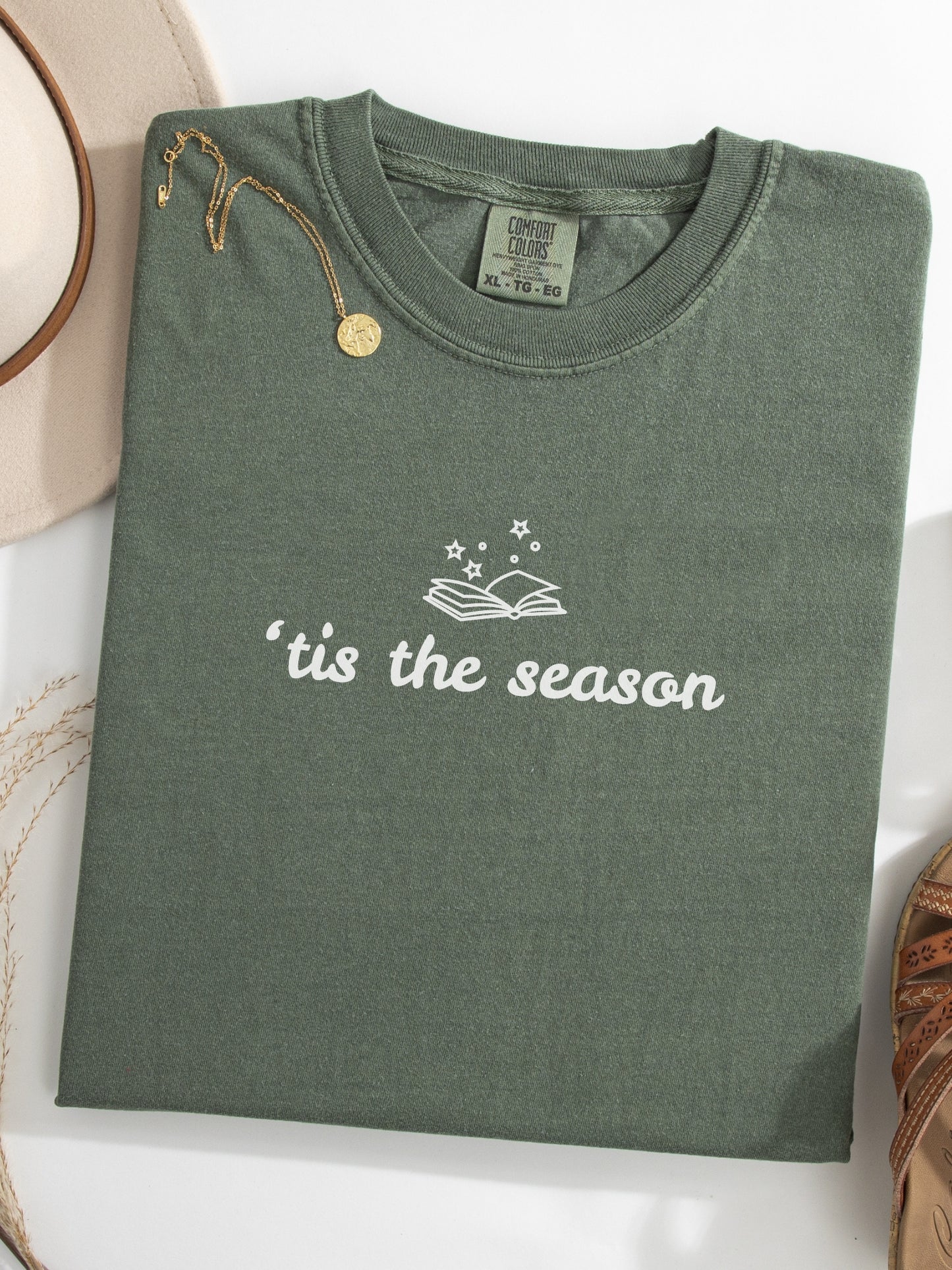 A moss green folded t-shirt with 'tis the season' printed in white on the front, styled with a gold pendant necklace and a beige hat on a white surface.