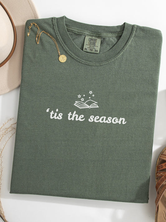 A moss green folded t-shirt with 'tis the season' printed in white on the front, styled with a gold pendant necklace and a beige hat on a white surface.
