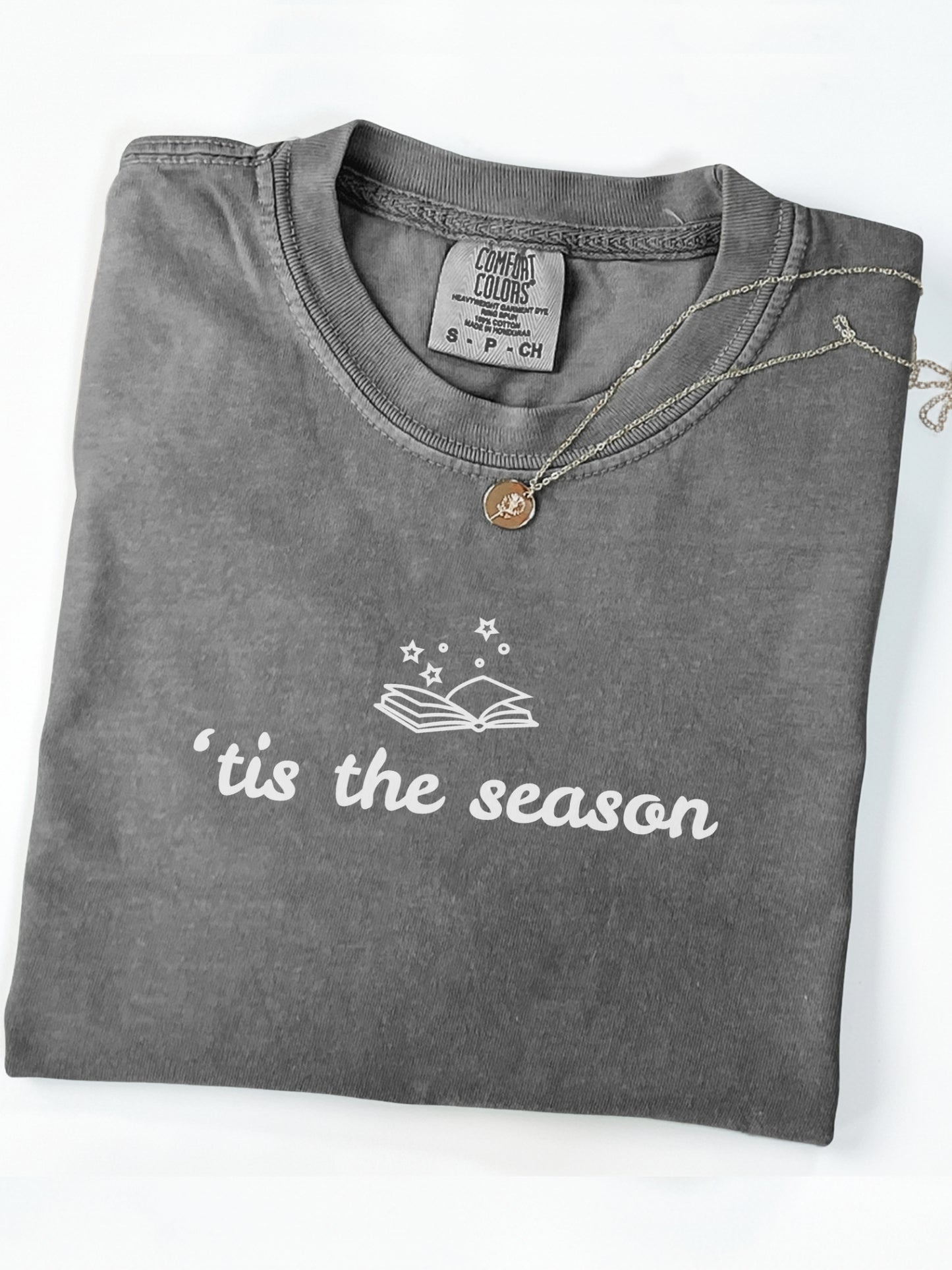 A folded pepper t-shirt with a white graphic print reading 'tis the season' and an open book with stars. A gold necklace with a letter 'A' pendant is draped over the collar.