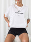A woman wearing a white short-sleeve crewneck t-shirt with a black graphic print that reads "'tis the season" below an open book illustration, paired with black shorts.