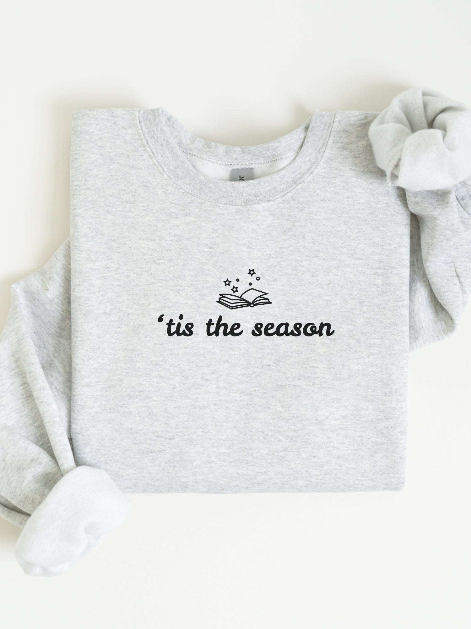 An ash heather gray sweatshirt laid flat on a white background, with the sleeves partially folded and a black graphic reading "'tis the season" beneath an open book and stars. A white fabric accessory is tucked into one sleeve.