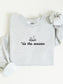 An ash heather gray sweatshirt laid flat on a white background, with the sleeves partially folded and a black graphic reading "'tis the season" beneath an open book and stars. A white fabric accessory is tucked into one sleeve.