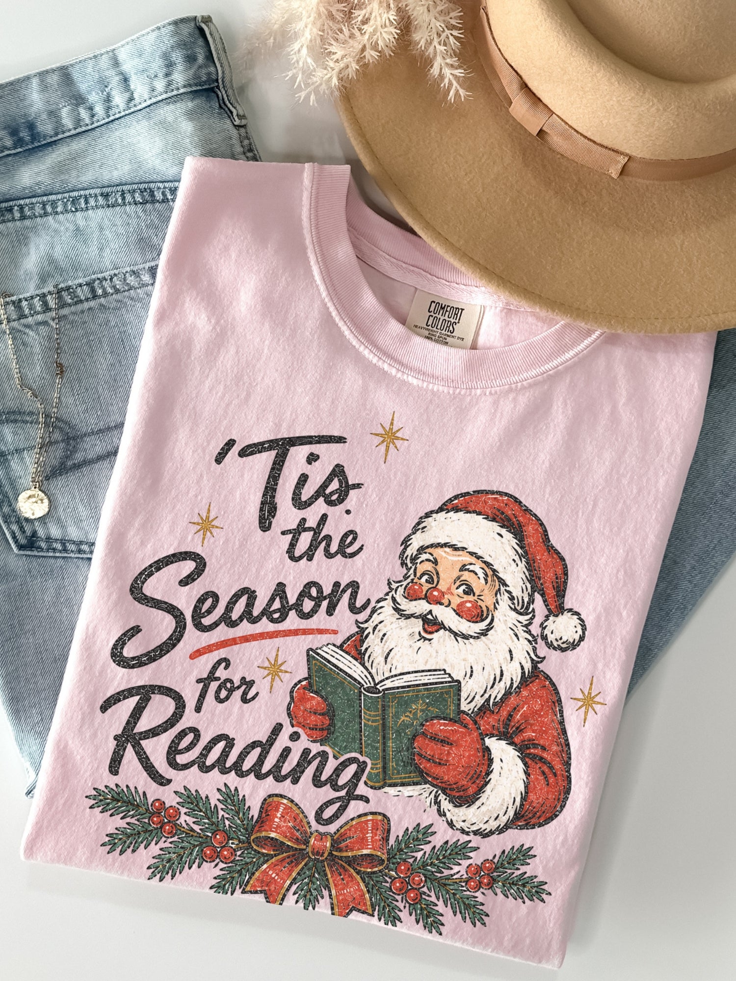 Blossom Comfort Colors shirt featuring a vintage Santa reading a green book with Tis the Season for Reading text, evergreen branches, red berries, festive bow and gold stars, Christmas book lover shirt