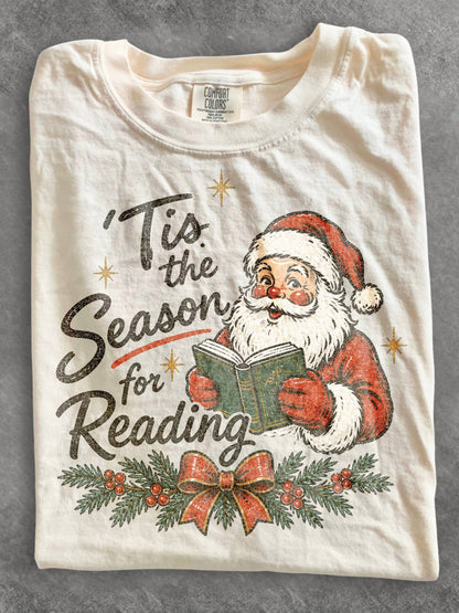 Ivory Comfort Colors shirt featuring a vintage Santa reading a book with Tis the Season for Reading design, evergreen branches, red berries, Christmas bow and gold stars, festive book lover Christmas shirt