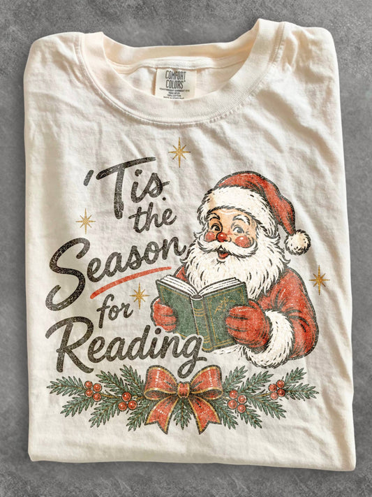 Ivory Comfort Colors shirt featuring a vintage Santa reading a book with Tis the Season for Reading design, evergreen branches, red berries, Christmas bow and gold stars, festive book lover Christmas shirt