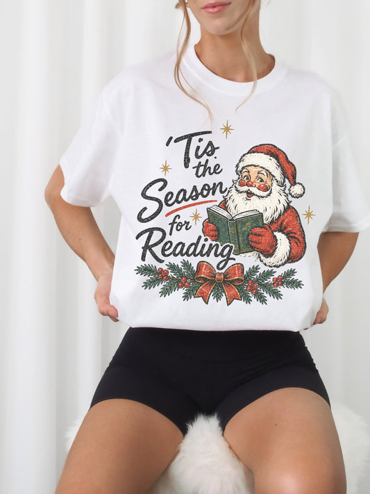 White Comfort Colors shirt modeled with a vintage Santa reading a green book and Tis the Season for Reading text, evergreen branches, red berries, festive bow and gold stars, Christmas reader shirt
