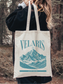 Canvas tote bag featuring a Velaris city of starlight mountain design from a fantasy inspired book theme, worn over the shoulder outdoors in a forest setting. The lifestyle image shows a cozy and adventurous aesthetic, perfect for fantasy readers and book lovers carrying books, groceries, or daily essentials.