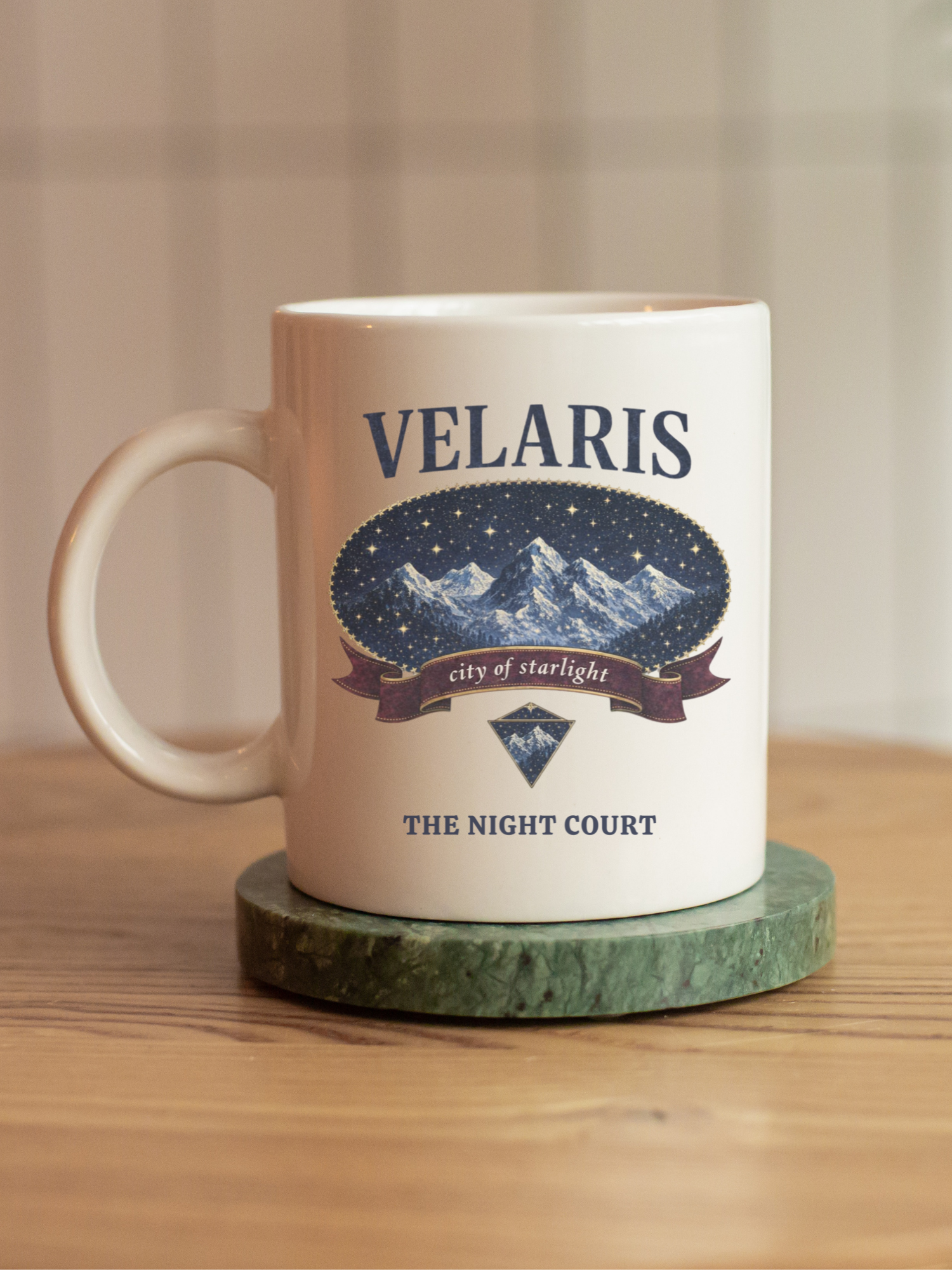 White ceramic mug featuring a Velaris city of starlight design from a fantasy inspired book theme, placed on a green marble coaster on a wooden table. The mug shows mountains and stars with night court text, styled in a cozy indoor setting with warm lighting, perfect for fantasy readers and book lovers who enjoy aesthetic drinkware.