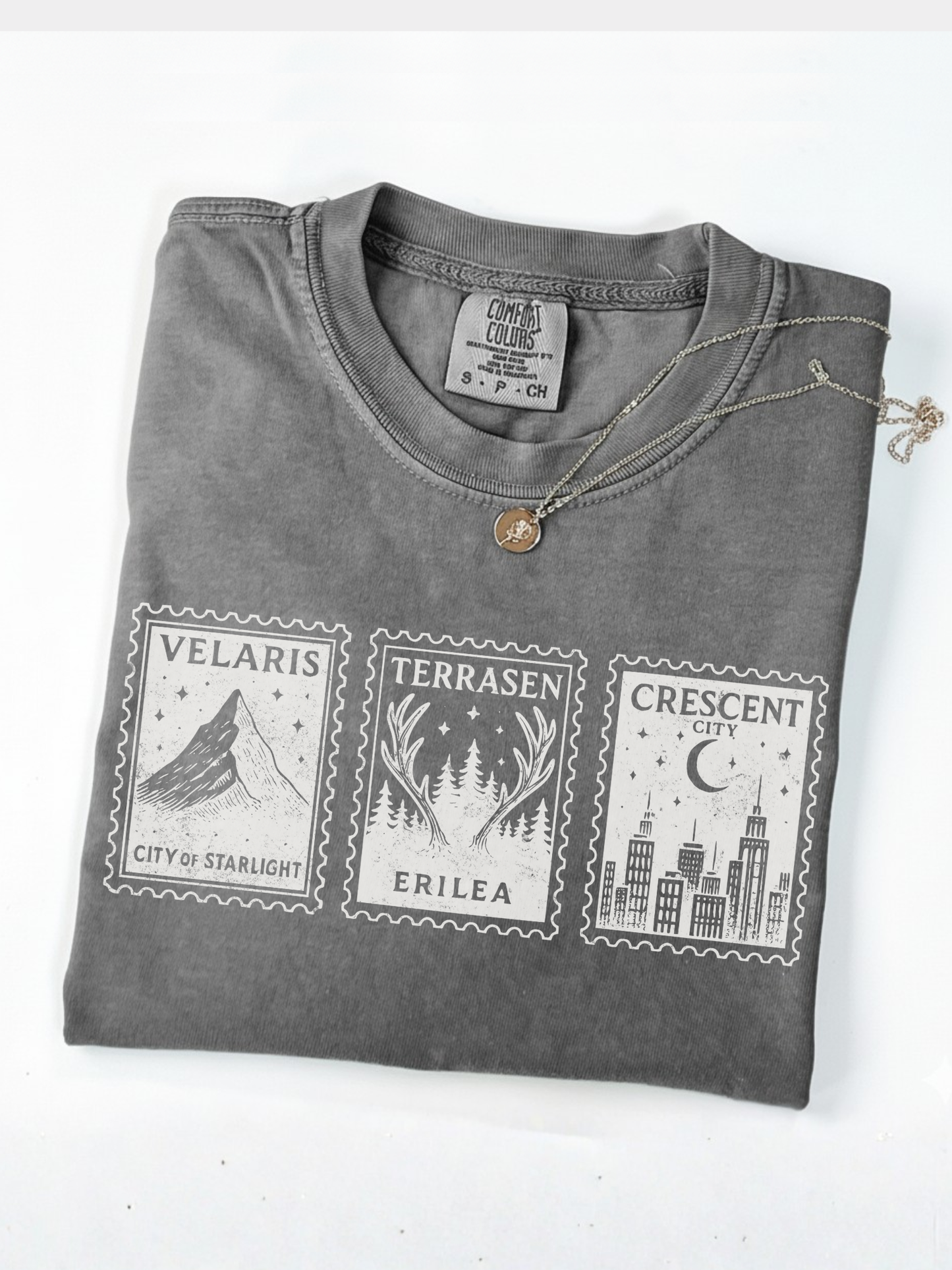 folded gray Comfort Colors shirt with Velaris, Terrasen, and Crescent City stamps next to gold necklace