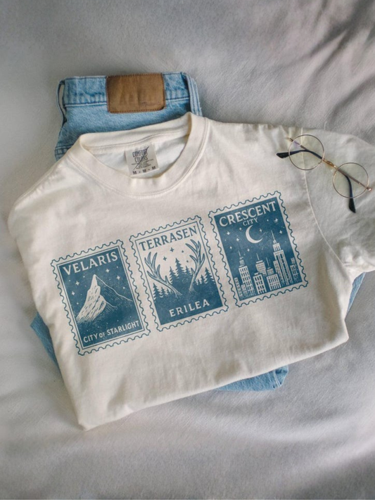 flat lay of folded Ivory Comfort Colors bookish t-shirt featuring Velaris, Terrasen, Crescent City stamps next to glasses and jeans