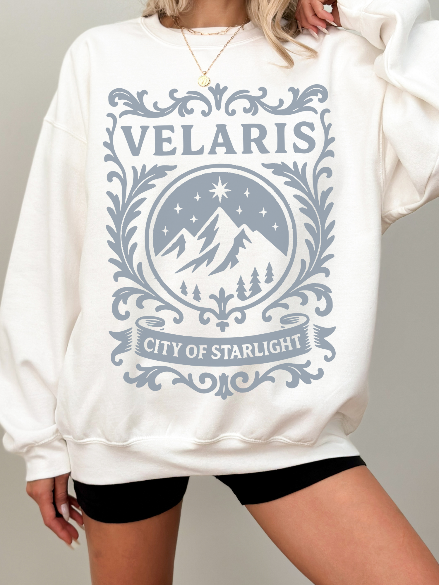woman wearing a white sweatshirt with soft grey Velaris City of Starlight design featuring stars, mountains, and scrollwork