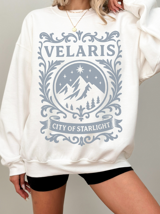 woman wearing a white sweatshirt with soft grey Velaris City of Starlight design featuring stars, mountains, and scrollwork