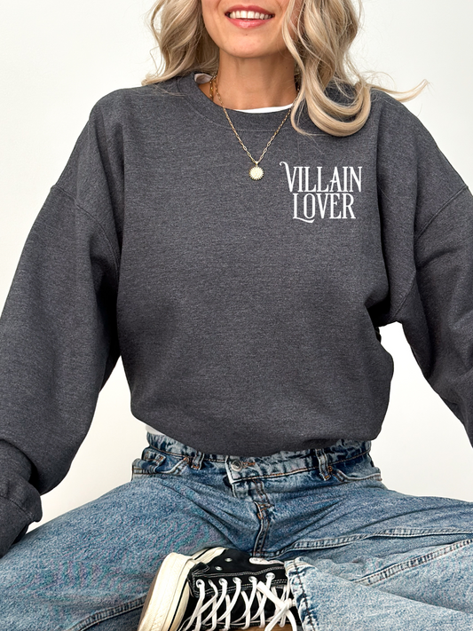 woman wearing dark heather sweatshirt with villain lover text, seated cross-legged in jeans and sneakers