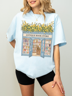 Woman modeling an oversized chambray t shirt with an antique bookstore graphic featuring bookshelves, open doors, and yellow flowers styled casually with black shorts