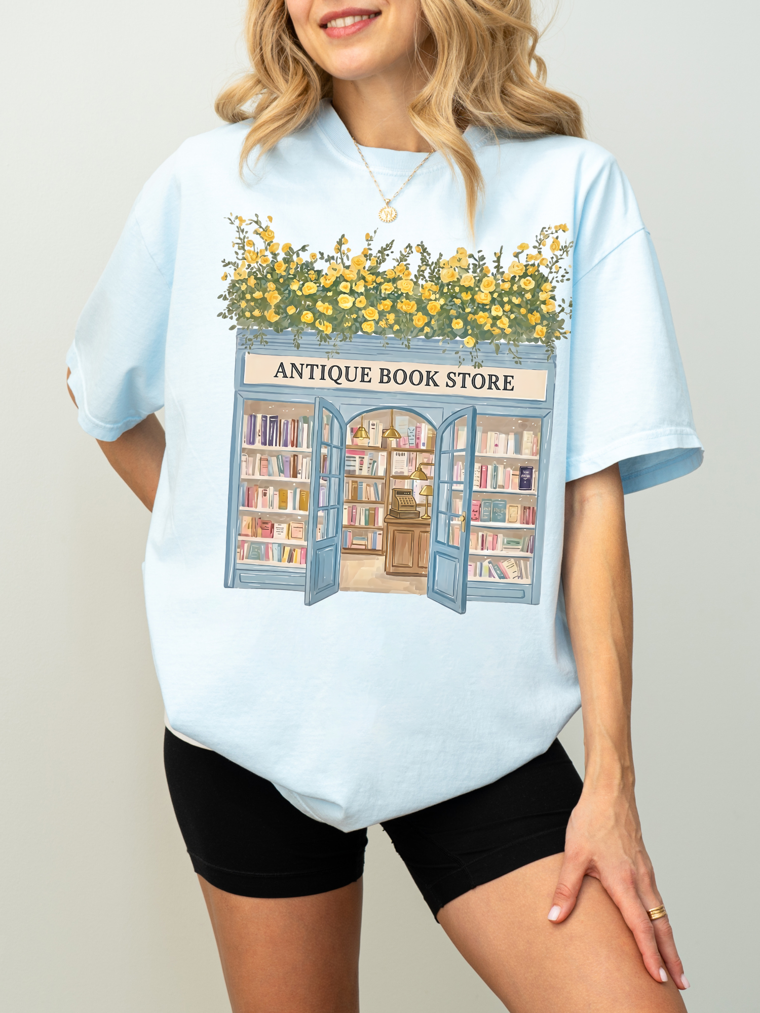 Woman modeling an oversized chambray t shirt with an antique bookstore graphic featuring bookshelves, open doors, and yellow flowers styled casually with black shorts