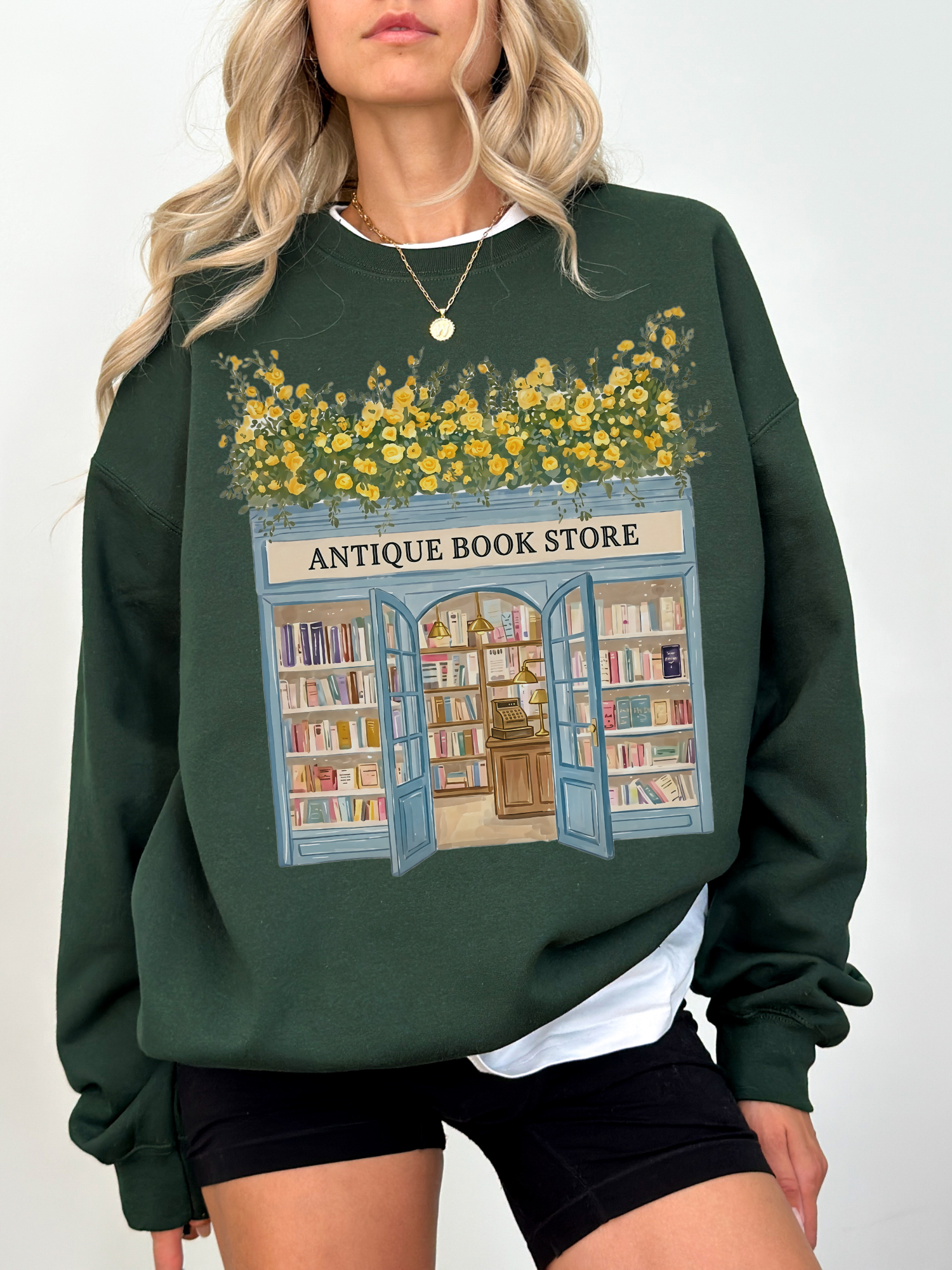 Woman wearing a dark green sweatshirt with an antique bookstore illustration featuring open doors, bookshelves, and yellow floral greenery styled in a casual outfit