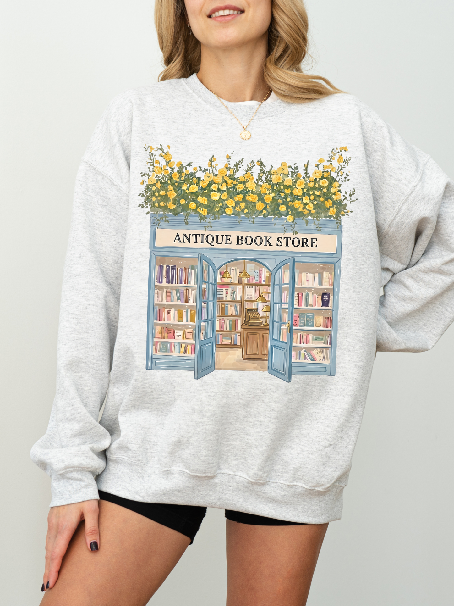 Woman modeling a gray sweatshirt with an antique bookstore design featuring bookshelves, open doors, and yellow flowers styled casually with a minimal background