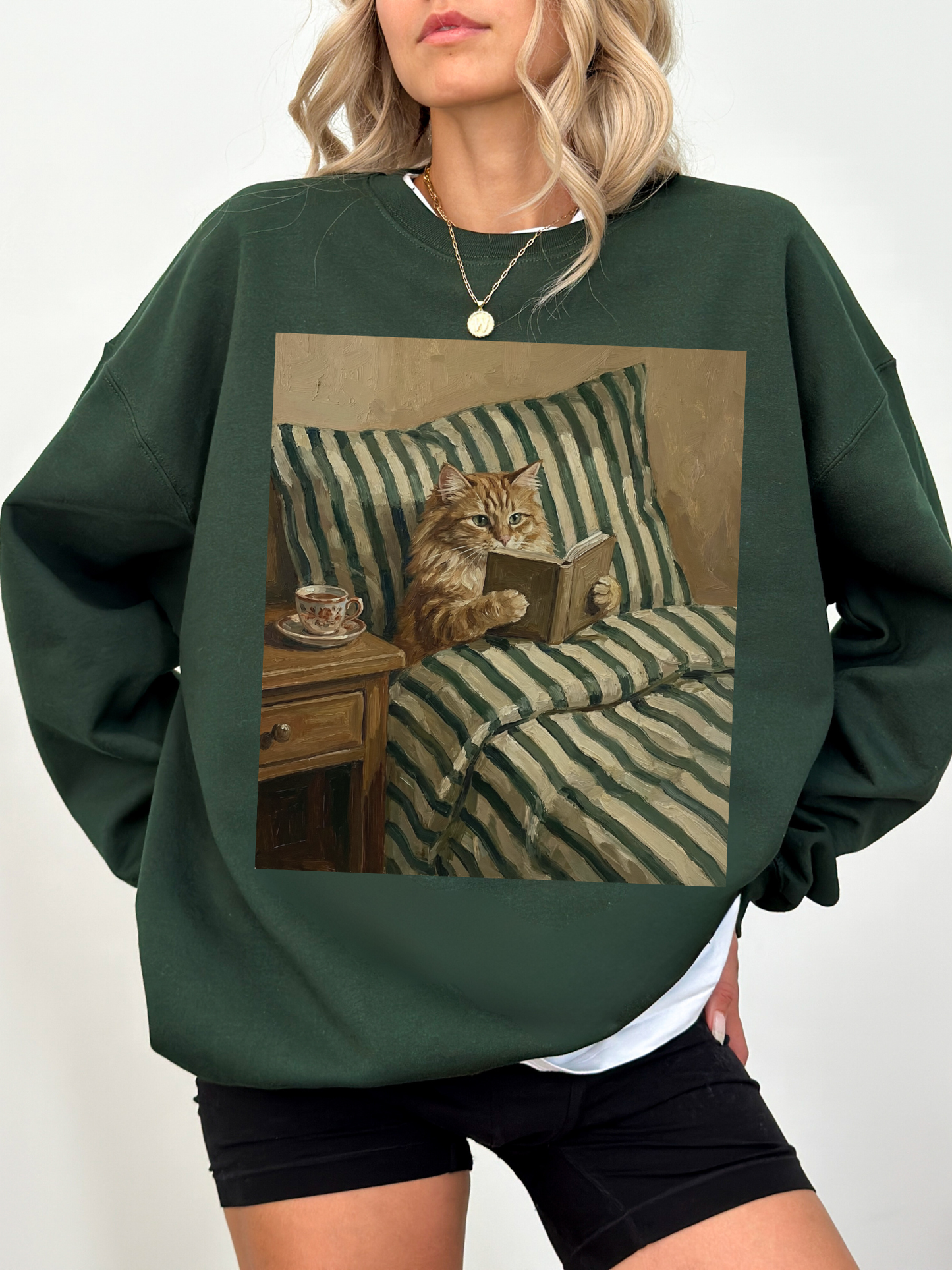 Dark green sweatshirt with a vintage inspired illustration of a cat reading a book in bed with striped blankets and a tea cup. Styled on a model indoors with layered jewelry, creating a cozy and aesthetic book lover look perfect for readers, cat lovers, and fall inspired outfits.