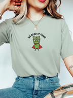 woman wearing a bay Comfort Colors shirt featuring a reading frog with a cape and We Read At Dawn design for book lovers
