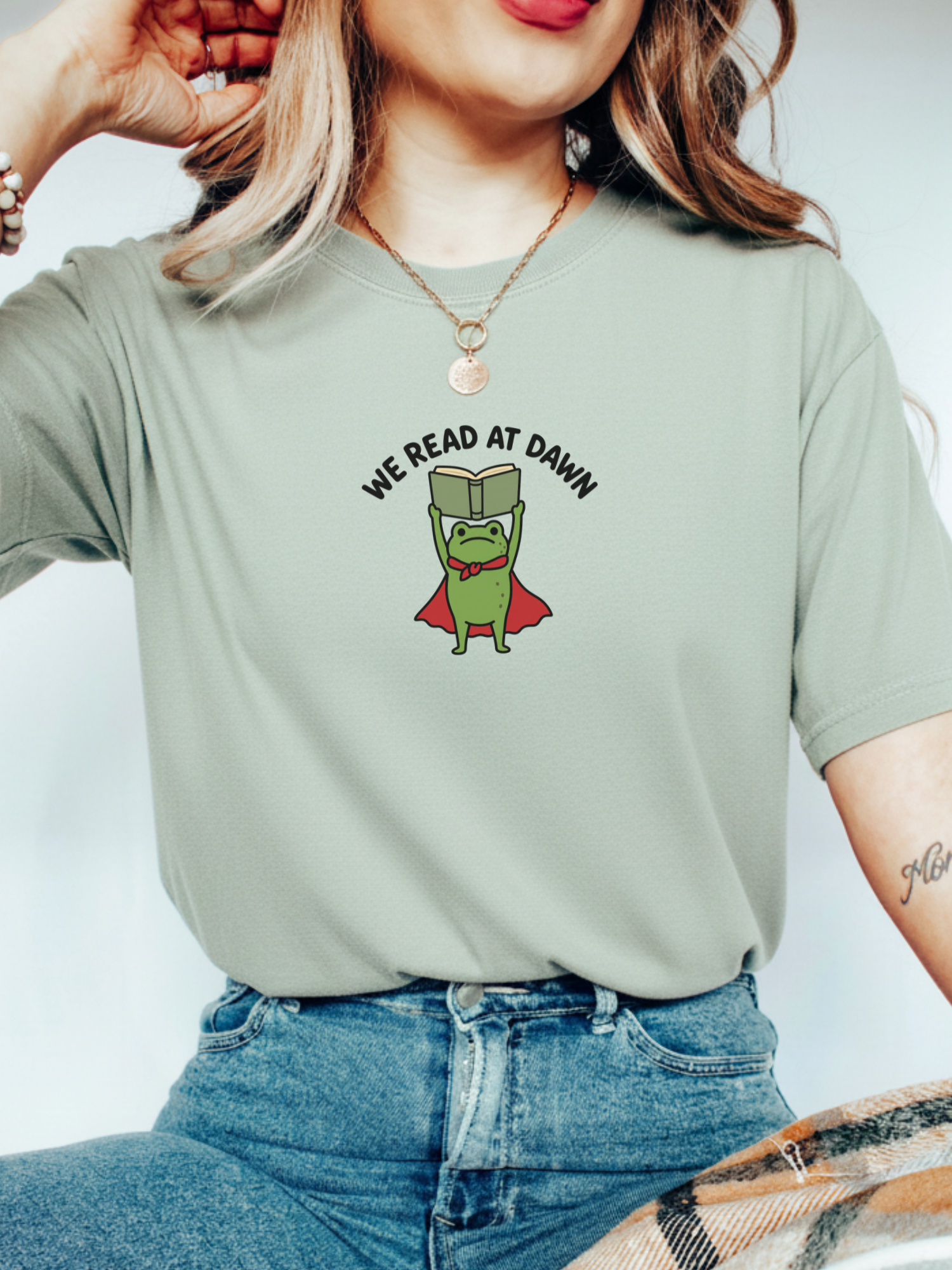 woman wearing a bay Comfort Colors shirt featuring a reading frog with a cape and We Read At Dawn design for book lovers