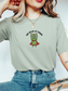 woman wearing a bay Comfort Colors shirt featuring a reading frog with a cape and We Read At Dawn design for book lovers