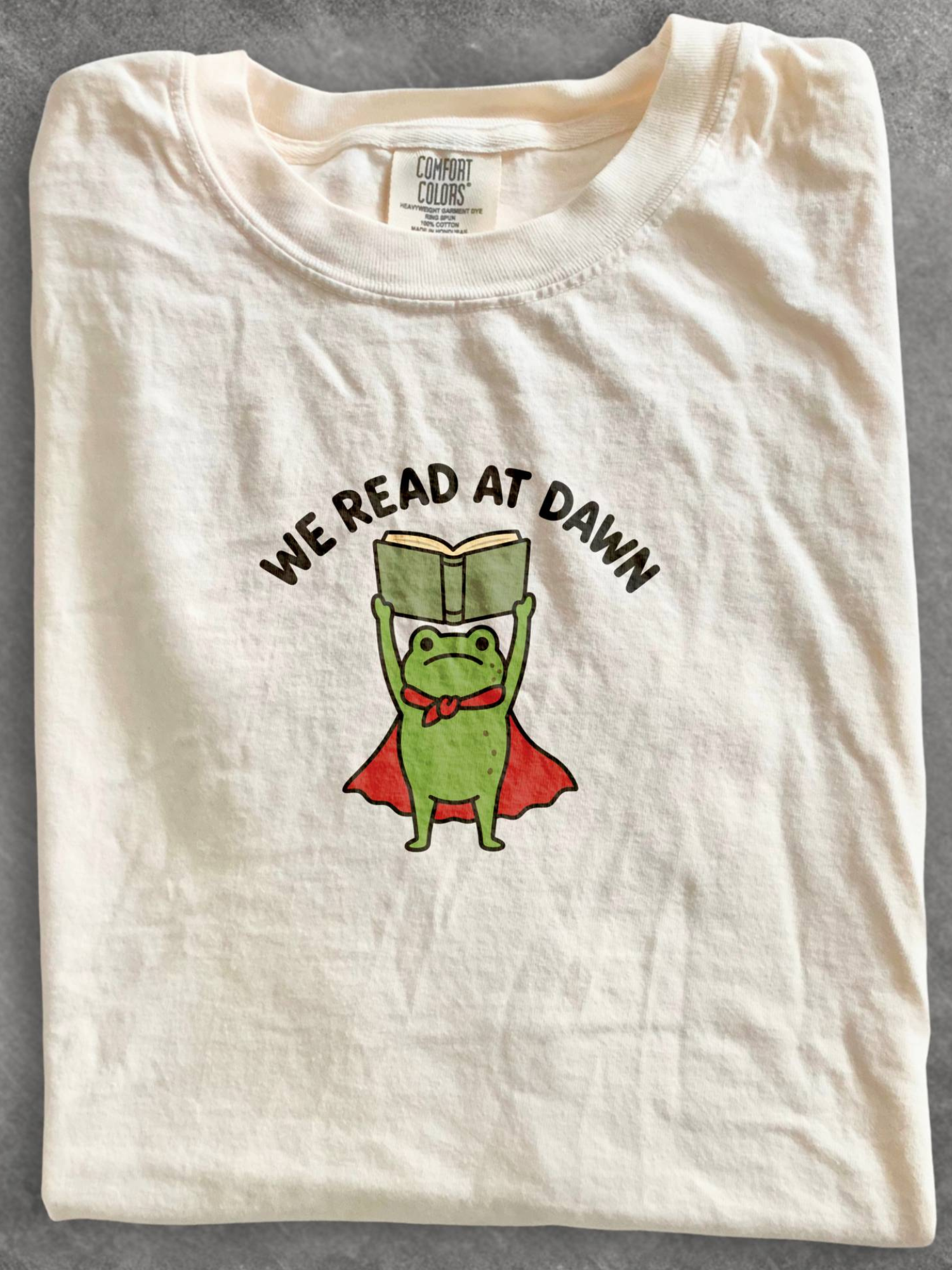 ivory Comfort Colors shirt flat lay featuring a reading frog with a cape and We Read At Dawn graphic for book lovers and readers