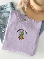 orchid Comfort Colors shirt flat lay featuring a reading frog with a superhero cape and We Read At Dawn book lover design