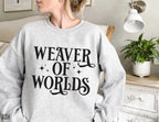 woman wearing Ash grey sweatshirt with “Weaver of Worlds” phrase in bold black text near white dresser
