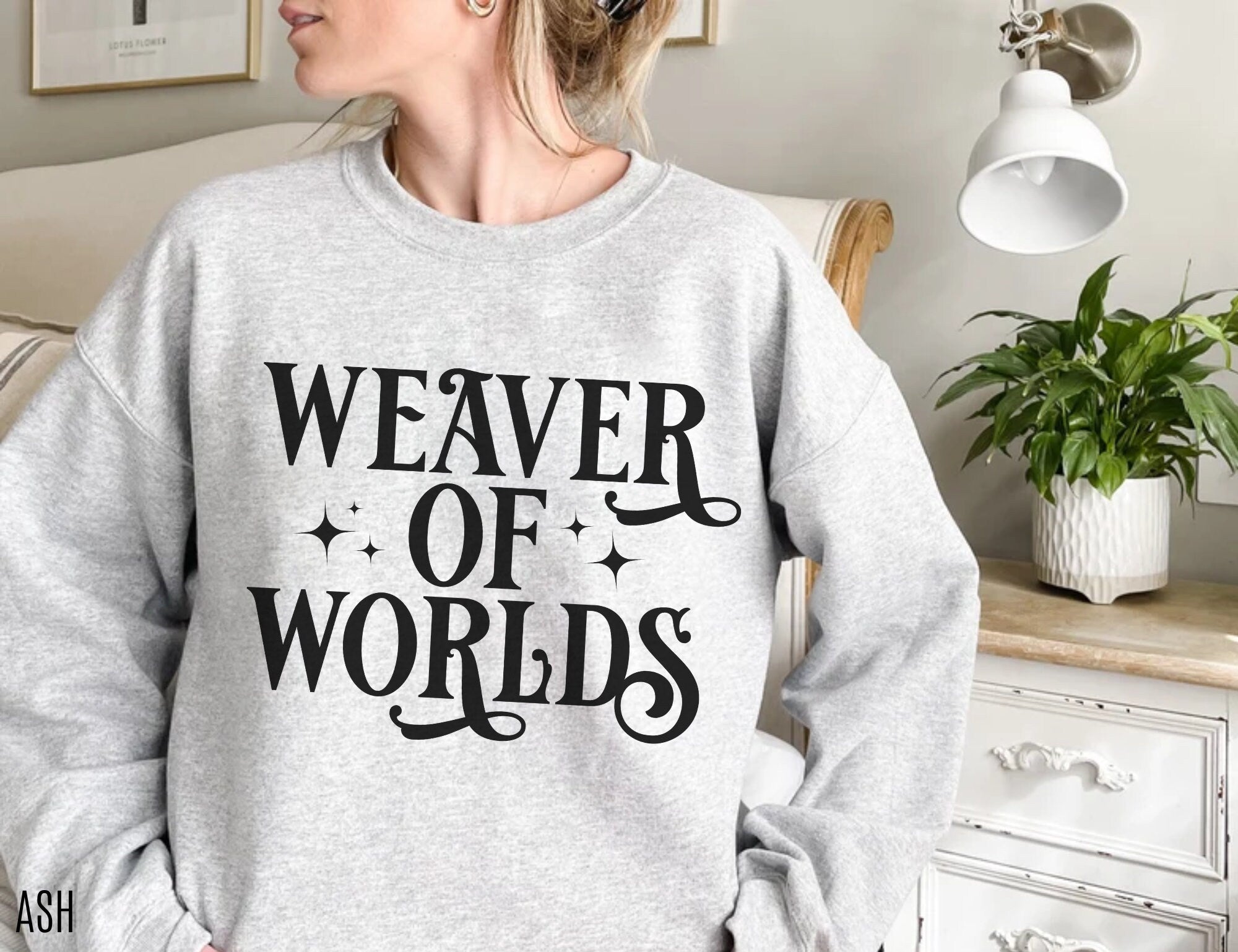 woman wearing Ash grey sweatshirt with “Weaver of Worlds” phrase in bold black text near white dresser