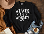folded black sweatshirt with white “Weaver of Worlds” print, laid out with jeans, hat, coffee, and a book
