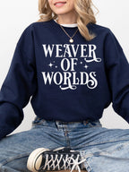 woman in navy blue sweatshirt with whimsical “Weaver of Worlds” design, sitting cross-legged in jeans