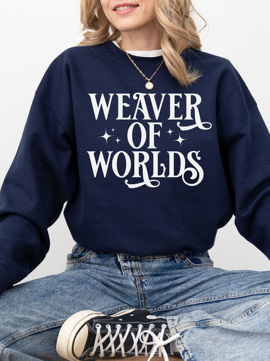 woman in navy blue sweatshirt with whimsical “Weaver of Worlds” design, sitting cross-legged in jeans