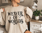 woman wearing sand-colored sweatshirt with black “Weaver of Worlds” text and sparkles beside a white nightstand