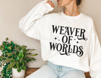 woman in white sweatshirt with fantasy-inspired “Weaver of Worlds” text and stars, sitting near green plants
