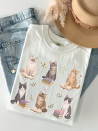 Flat lay of white Comfort Colors t shirt featuring cats reading books with floral accents styled with denim jeans gold necklace and beige hat