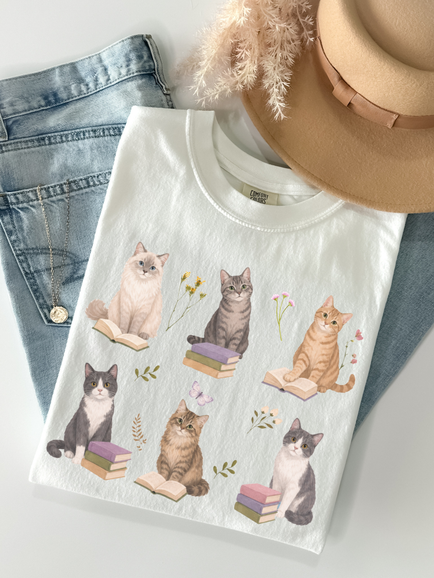 Flat lay of white Comfort Colors t shirt featuring cats reading books with floral accents styled with denim jeans gold necklace and beige hat