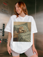 White oversized t-shirt with a vintage style cat reading a book illustration, styled on a model inside an elevator with casual lounge shorts. Modern lifestyle photo combining cozy reading vibes with everyday street style for book lovers and cat lovers.