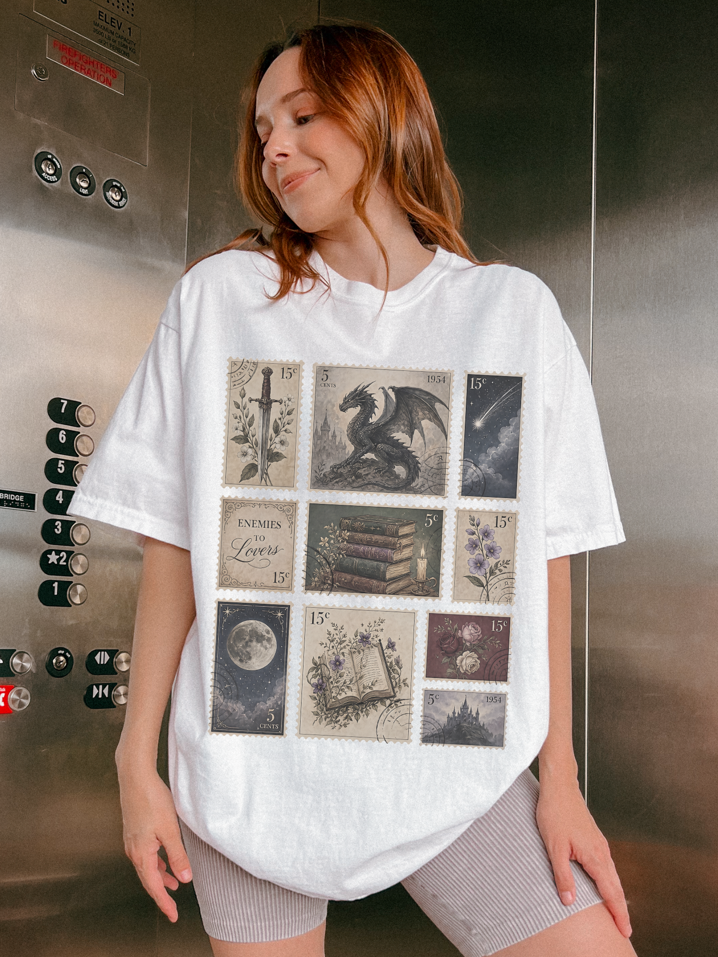 Woman wearing oversized white fantasy bookish t shirt featuring vintage postage stamp collage with dragon sword moon books flowers castle and enemies to lovers artwork inside an elevator