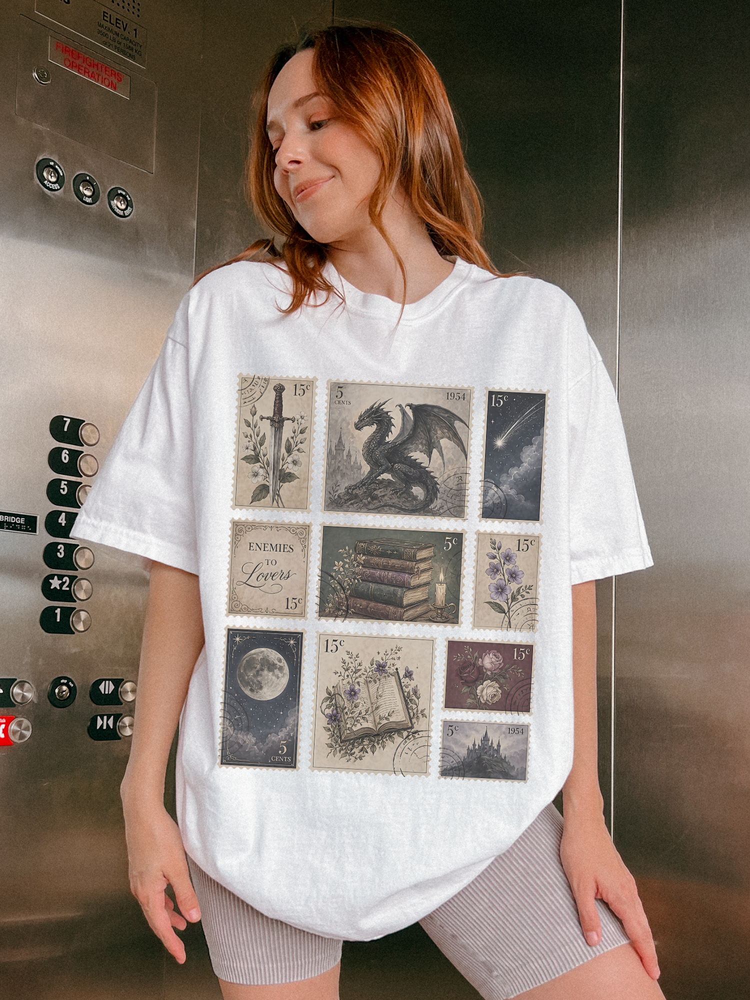 Woman wearing oversized white fantasy bookish t shirt featuring vintage postage stamp collage with dragon sword moon books flowers castle and enemies to lovers artwork inside an elevator