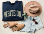 flatlay of folded navy write on sweatshirt with distressed cream text, paired with shorts, hat, and sunglasses