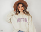smiling woman in sand-colored sweatshirt with lavender write on text and wide-brim hat
