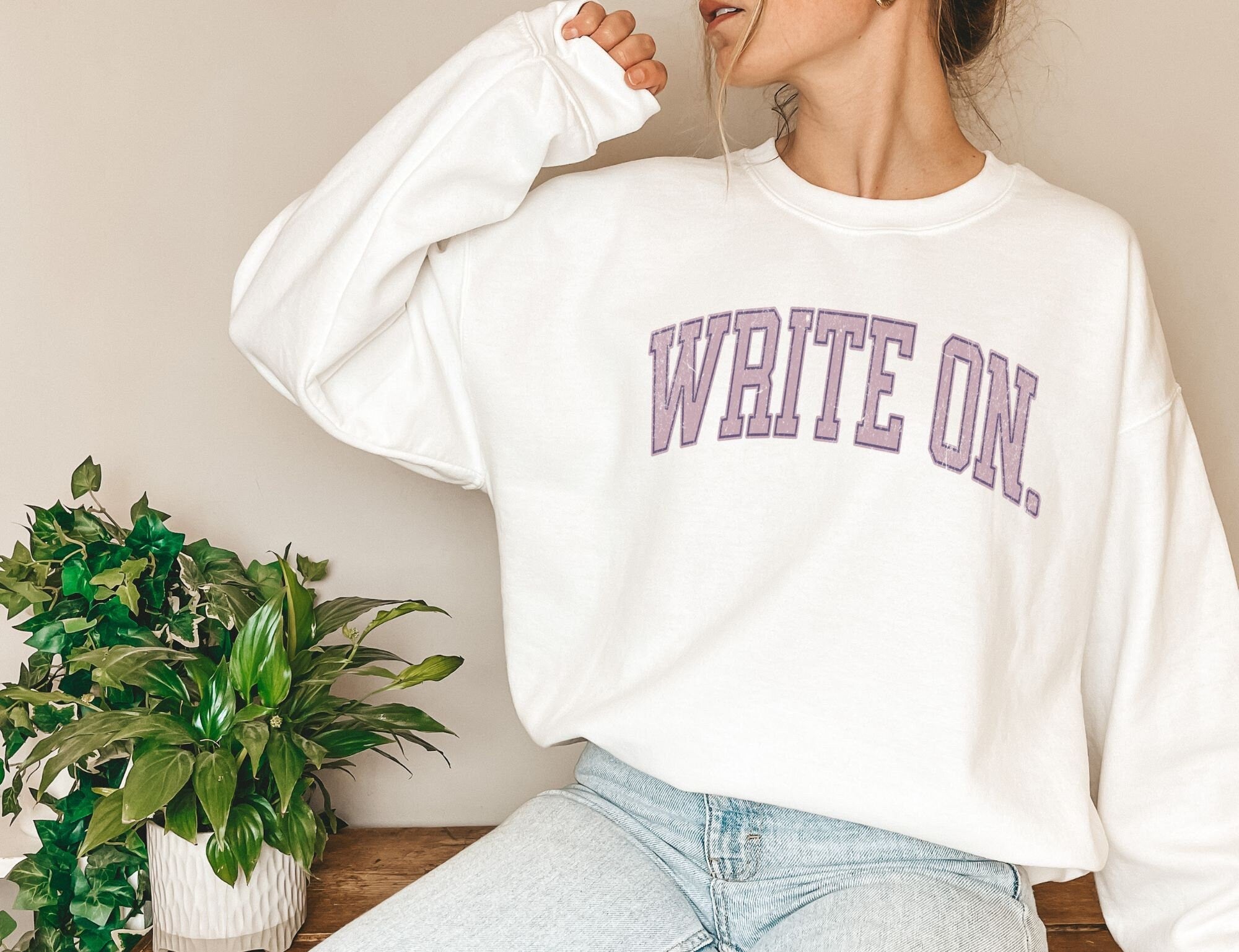 woman in white sweatshirt with purple write on text and light denim jeans, sitting beside plant
